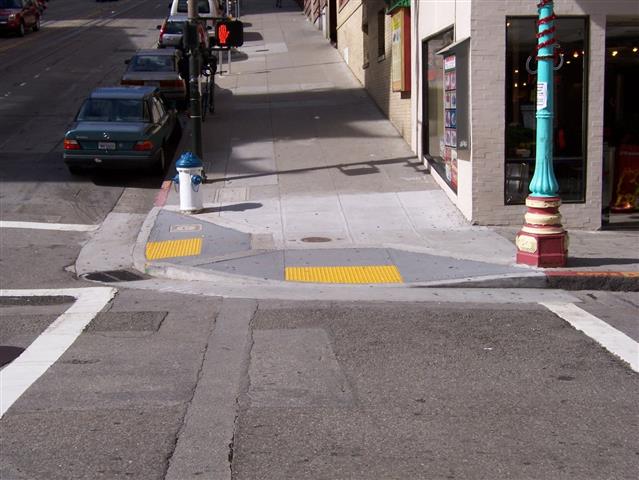 Attention TWSIs used on curb ramps at an intersection. The TWSIs are bright yellow, in contrast to the adjacent concrete and asphalt ground finishes.